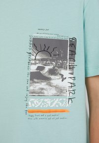 Licht turquoise katoenen t-shirt met een zwart grafisch ontwerp van een skateboard scène, zon en tekstaccenten in verschillende lettertypen.