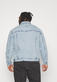 Vaaleansininen farkkutakki, jossa on klassinen kaulus, pitkät hihat ja kontrastitimanttisaumat. Napitus hihansuissa ja takana oleva kaarroke yksityiskohtana.