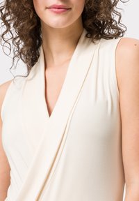 Blusa sin mangas beige con diseño envolvente, confeccionada en una tela suave con una textura lisa y escote drapeado.