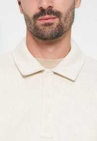 Gros plan d’un homme avec une barbe courte et foncée portant un polo crème boutonné avec un col plié.