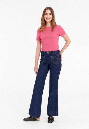 Chemise rose en coton côtelé à manches courtes associée à un jean évasé bleu foncé avec poches avant et couture contrastante ; modèle se tenant devant un fond neutre.