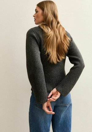 Donkergrijze wollen trui met lange, wijde mouwen, gecombineerd met blauwe spijkerbroek. Haar stijl recht, van achteren gezien.