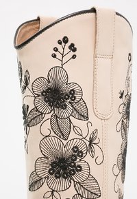 Stivale in pelle crema alto fino al ginocchio, caratterizzato da un ricamo floreale nero dettagliato, una finitura liscia e un'apertura stretta con una cucitura.