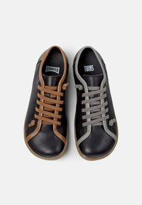 Schwarze Schuhe mit kontrastierenden Schnürsenkeln; einer hat braune Schnürsenkel mit dem Aufdruck "CAMPER," der andere graue Schnürsenkel mit dem Aufdruck "TWINS." Einfaches, modernes Design.