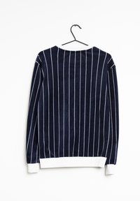 Marineblauwe top met lange mouwen en verticale witte strepen, textuurrijke stof, ronde halslijn, en witte tailleband en manchetten. Hangt aan een zwarte haak.