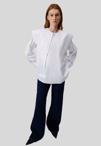 Donna con capelli rossi lisci che indossa una blusa bianca con un ampio colletto triangolare e pantaloni a gamba larga scuri su uno sfondo grigio.