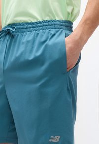 Άνδρας που φοράει πετρόλ σορτς New Balance με ελαστική μέση και κορδόνι, με το χέρι στην δεξιά τσέπη, συνδυασμένο με ανοιχτό πράσινο πουκάμισο.