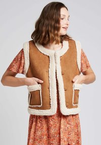 Gilet en suédine marron avec un col arrondi et un revêtement en fausse fourrure crème, doté de deux poches plaquées à l'avant. Porté par-dessus une robe à fleurs.