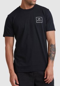 Zwarte katoenen t-shirt met korte mouwen, een ronde hals en een wit rechthoekig logo op de linkerkant van de borst. Gladde textuur, relaxte pasvorm.