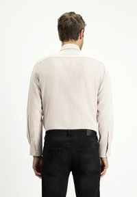 Camicia a maniche lunghe beige chiaro con polsini abbottonati e taglio dritto, abbinata a jeans neri. Texture liscia, senza motivi né accenti.