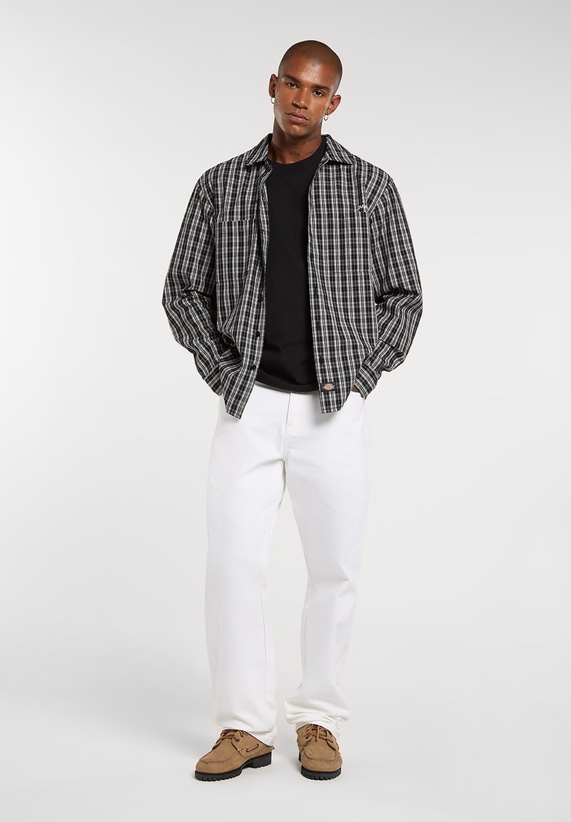 Camicia a quadri nera, bianca e grigia sopra una t-shirt nera, abbinata a pantaloni bianchi e scarpe marroni. Vestibilità casual, design con bottoni frontali.