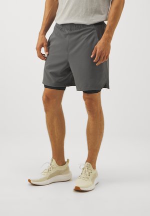 Male wearing gray athletic shorts with black inner layer and beige running shoes, standing against white background.