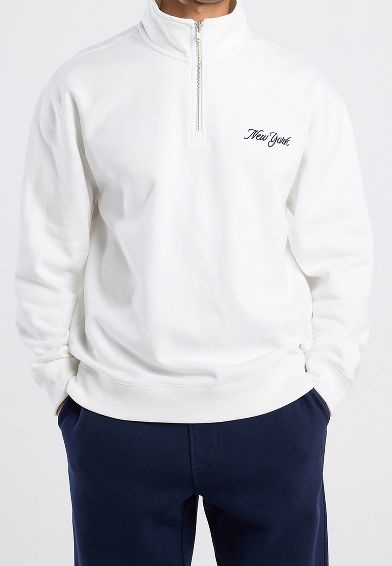 Homme portant un sweat-shirt blanc à zip court avec le texte "New York" sur la poitrine et un pantalon bleu foncé, mains dans les poches.
