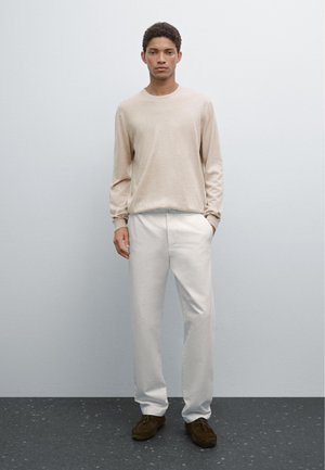 Light beige long-sleeve shirt with a crew neckline, paired with white trousers. Model stands on a dark textured surface. Brown shoes.