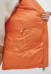 Oranje geblokte jas interieur met een pocket met een tab, zilveren drukknoopjes en een cirkelvormige waslabelgrafiek. Glad, lichtgewicht textuur.