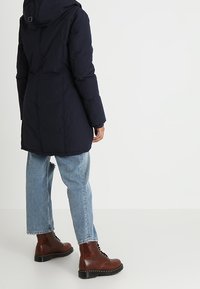 Mörkblå pufferjacka med huva, med quiltat mönster och långa ärmar, i kombination med ljusblå jeans och bruna snörstövlar.