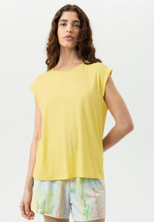 Femme aux cheveux bruns ondulés portant un haut jaune sans manches ample et un short pastel multicolore à motifs, sur fond uni.