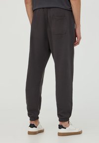 Muž v tmavě šedých teplácích s kapsou na zadní straně a bílých teniskách s černými detaily na patách, stojící na jednobarevném pozadí.