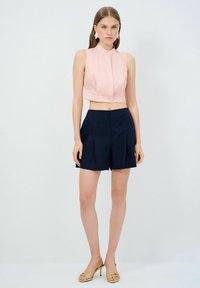 Blusa corta rosa con colletto alla mandarina e chiusura a bottone, abbinata a pantaloni corti blu navy a vita alta con pieghe. Sandali dorati completano il look.
