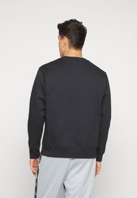 Hombre de espaldas que lleva una sudadera negra de manga larga y pantalones gris claro con franjas laterales en negro y blanco.