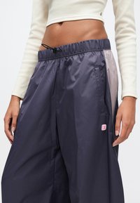 Μεσαίο τμήμα ατόμου που φοράει φαρδιά συρραπτόμενα παντελόνια από σκούρο μωβ αδιάβροχο υλικό με ελαστική μέση και πλαϊνές τσέπες, σε συνδυασμό με κομμένο κρεμ ριμπ τοπ.