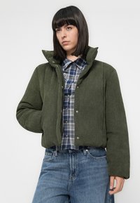 Vrouw met steil, donker haar in een olijfgroene gewatteerde jas, een blauw geblokt overhemd met knopen en blauwe spijkerbroek tegen een witte achtergrond.