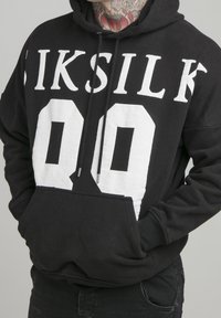 Černá mikina s velkým bílým nápisem "SIKSILK 00" na přední straně. Má kapsu na přední straně a stahovací šňůrky s kovovými koncovkami. Texturovaná látka.