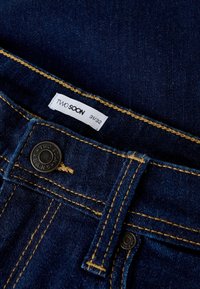 Tamsiai mėlynos spalvos džinsai su geltonais siūlais. Yra juodas metalinis mygtukas ir baltas etiketė su užrašu „TWO SOON 31/32.“