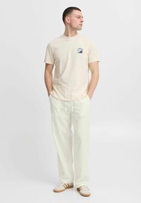 Beige katoenen t-shirt met een kleine blauwe grafiek op de borst, gecombineerd met losse witte broeken en sneakers met bruine accenten.