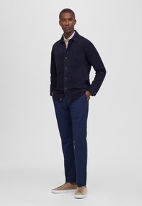 Chaqueta de ante azul marino con cierre de botones, dos bolsillos en el pecho y cuello con solapa. Combinada con pantalones de cuadros azul marino y zapatillas beige.