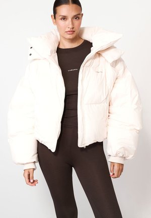 Puchowa biała kurtka z wysokim kołnierzem, na suwak i z bocznymi kieszeniami; ma ściągacze przy mankietach oraz gładką, śliską teksturę. Nosi się ją na brązowej bluzce.