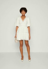 Λευκό, κοντό φόρεμα με φουσκωτά μανίκια, κομψό ντεκολτέ και λεπτομέρειες από δαντέλα. Φορεμένο με λευκά ψηλοτάκουνα και κρατώντας ένα κρεμ χρώμα clutch.