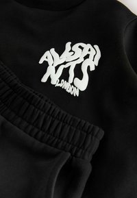 Čierna mikina s bielym grafickým textom "ALVSANTS LONDON." Má rebrovaný kulatý výstrih a elastické detaily na manžetách. Hladká textúra látky.