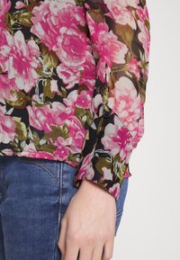 Blusa floreale in tessuto nero trasparente con motivo floreale rosa e verde, dotata di polsini con bottone e una vestibilità rilassata.