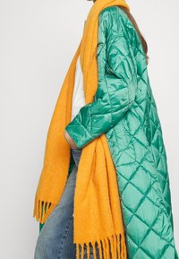 Cappotto trapuntato verde con una finitura lucida, abbinato a una sciarpa di lana arancione brillante con frange. Camicia bianca casual e denim visibili.