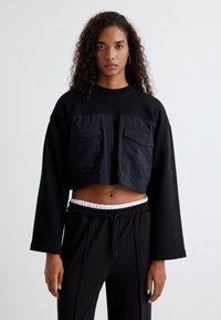 Schwarzer, kurz geschnittener Sweatshirt mit langen Ärmeln, ausgestattet mit einem strukturierten Frontpanel und zwei Brusttaschen. Kombiniert mit schwarzen Hosen und einem weißen Bund.