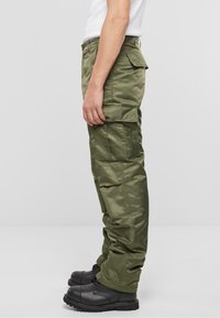 Pantalons cargo vert olive avec une finition lisse et brillante, dotés de plusieurs poches, d'une coupe décontractée et de bottes à lacets noires.