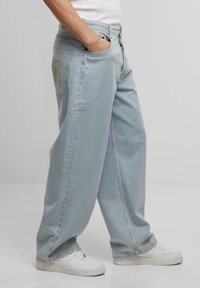 Lyseblå vide jeans laget av denim, med frontlommer, knappestolpe og en stor brodert logo på baklommen.
