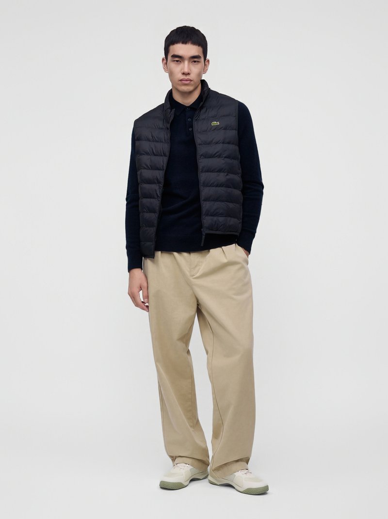 Jeune homme portant une veste matelassée noire, une chemise noire à manches longues, un pantalon beige à jambes larges et des baskets blanches, debout devant un fond uni.