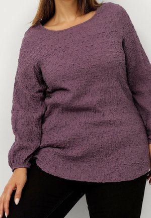 Personne portant un pull violet texturé à manches longues et un pantalon noir, avec une main posée sur la cuisse.