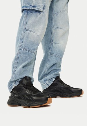 Osoba nosząca jasnoniebieskie dżinsy i czarne trampki Reebok z grubą podeszwą oraz gumową podeszwą zewnętrzną.