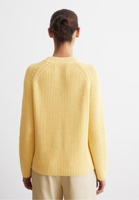 Donna con i capelli castani raccolti a chignon, che indossa un maglione giallo a coste e pantaloni beige, mostrata di spalle su uno sfondo bianco.