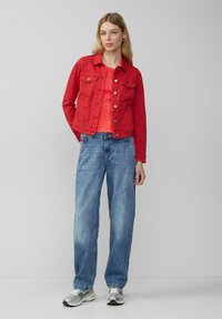Rode spijkerjack met zilveren knopen, gecombineerd met een rode top en lichtblauwe wijde jeans. Schoenen hebben een zilveren afwerking en een gaasstructuur.