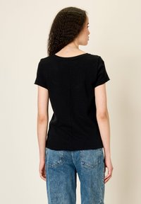 Žena s kudrnatými vlasy, oblečená v černém tričku s krátkým rukávem a modrých džínách, stojící zády k fotoaparátu před jednobarevným pozadím.