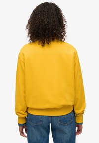 Felpa gialla con polsini e orlo a coste, realizzata con una vestibilità rilassata. Abbinata a jeans in denim blu, che mostrano uno stile casual.