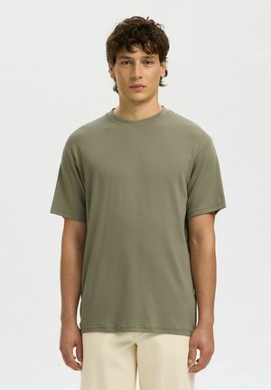 Jeune homme aux cheveux bruns bouclés portant un t-shirt vert olive uni à manches courtes et un pantalon beige, debout devant un fond blanc.