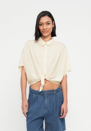 Kurzärmeliges, cremefarbenes Hemd mit Knoten vorne, Knopfdetails und lockerem Sitz, kombiniert mit blauen Shorts mit Nadelstreifen.