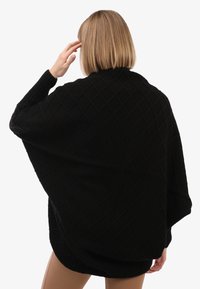 Blond bobikärniga naine mustas tekstuuriga kudumisjakus, seismas kaamera selja poole, käsi pea lähedal.