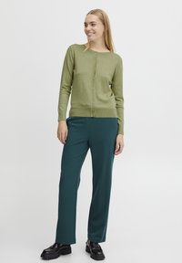 Cardigan vert à manches longues avec fermeture à boutons, associé à un pantalon sarouel teal. Des mocassins noirs complètent la tenue. Texture lisse dans l'ensemble.