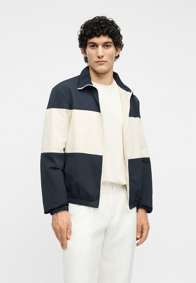 Νεαρός άνδρας με κοντά σγουρά μαύρα μαλλιά που φοράει μπλε σκούρο και εκρού μπουφάν με φερμουάρ και λευκό παντελόνι σε απλό φόντο.